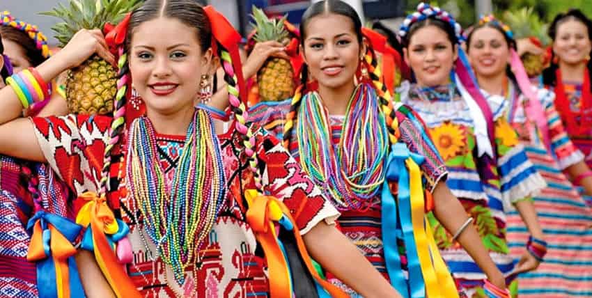 Un grupo de jóvenes mujeres mexicanas sonrientes, adornadas con una vibrante atuendo tradicional de oaxaca indígena, participan en una celebración cultural. En primer plano, dos mujeres con cabello intrincadamente trenzado decorado con cintas coloridas usan blusas y collares ricamente bordados hechos de innumerables hilos de cuentas multicolores. Algunas de las mujeres sostienen una piña en el hombro derecho. Otras mujeres con ropa festiva similar están borrosas en el fondo, lo que sugiere un desfile o baile.