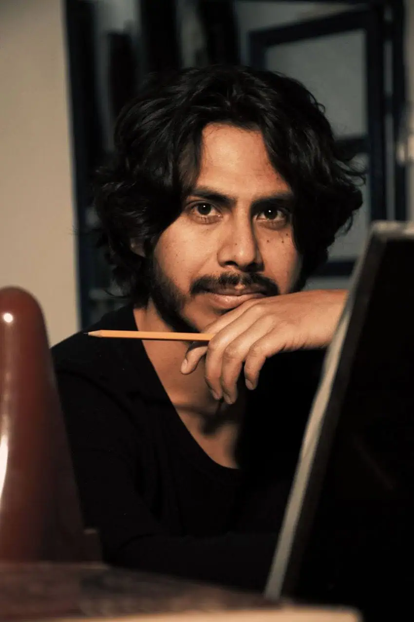 Un hombre con cabello ondulado oscuro y una perilla se ve considerablemente en la cámara. Está descansando la barbilla en su mano y sosteniendo un lápiz amarillo, sentado detrás de un caballete en un entorno artístico y cálidamente iluminado.