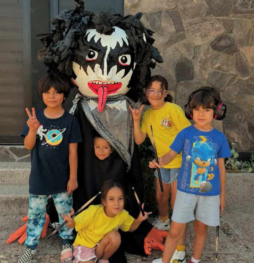 Cinco niños pequeños posan al aire libre frente a un muro de piedra con un gran títere mexicano de mojiganga hecho para parecer un miembro de la banda de rock beso en maquillaje de cara en blanco y negro y una lengua larga. Los niños están sonriendo a su alrededor, algunos sosteniendo baquetas y otros que hacen letreros manuales de paz o "rock-on".