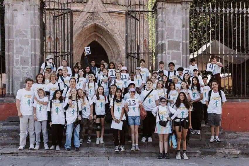 Un gran grupo de adolescentes y adultos jóvenes posa para una fotografía grupal en los escalones de piedra frente a una iglesia histórica e adornada en San Miguel de Allende después de la carrera anual de la ciudad de la ciudad. La mayoría usa camisetas blancas a juego con un logotipo, y varias están sosteniendo señales con números que parecen ser sus pancartas correctas.