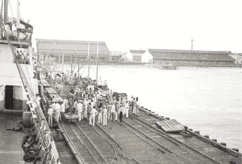 Foto en blanco y negro de un concurrido escenario de muelle que se viste con ropa blanca y sombreros, parados en un muelle de madera al lado de un gran barco en México. Algunos están empujando carros de plataforma en vías del ferrocarril que corren a lo largo del muelle. En el fondo, hay un gran cuerpo de agua y varios edificios de aspecto industrial.