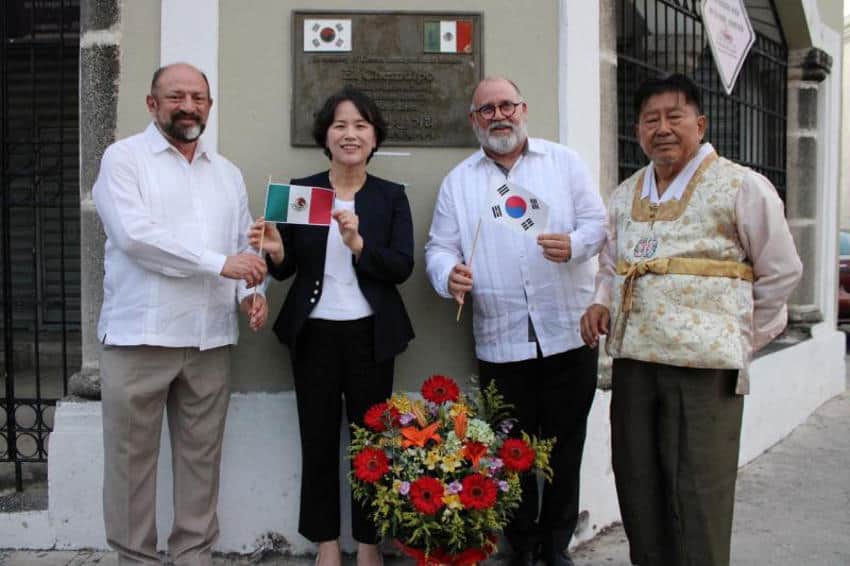 Una foto en color de cuatro funcionarios gubernamentales de Corea y México que oponen al aire libre en Mérida, México, frente a una placa de bronce. La persona en el extremo izquierdo y el segundo de la derecha son hombres que sostienen pequeñas banderas mexicanas y surcoreanas. Están vestidos con camisas blancas y pantalones de color claro. La mujer en el centro sostiene una bandera mexicana y está vestida con una blusa oscura y una blusa blanca. El hombre de la extrema derecha usa ropa coreana tradicional, incluido un chaleco de estilo Hanbok. Una corona de flores se encuentra en el suelo frente al grupo.