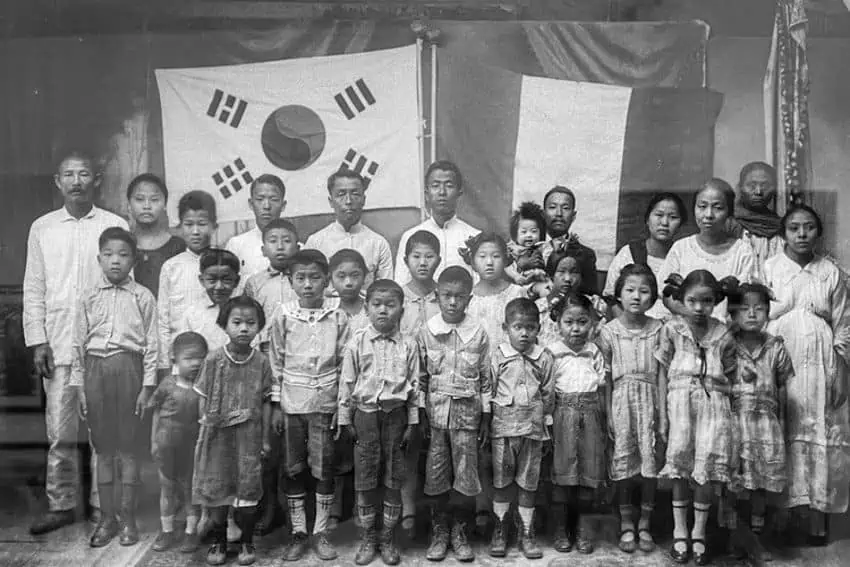 Black and white photo of a group of Mexican Koreans and Maya people posing together in front of a wall bearing two flags — one Korean and another a banner reminiscent of the Mexican flag. The group includes several adults and many children. The adults are standing in the back row while the children are in front. The subjects are dressed in simple, everyday clothing.