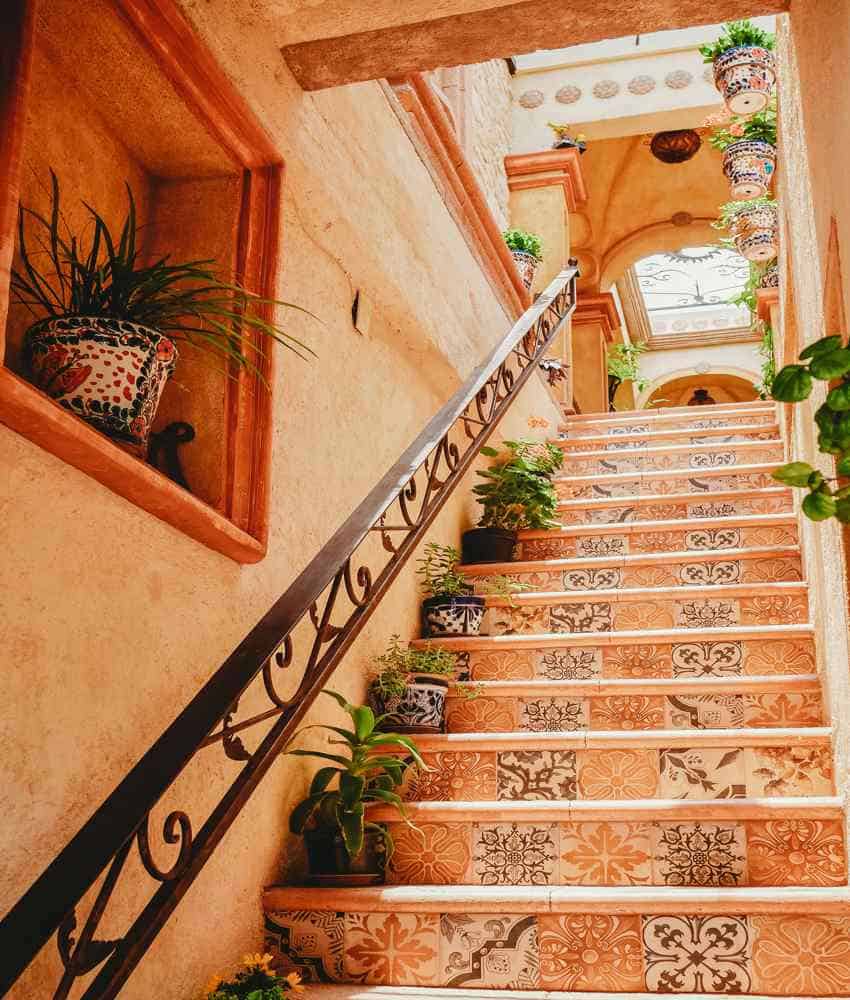 Una colorida escalera en México con cada paso decorada con un azulejo único y estampado, que conduce a un pasillo de segundo piso iluminado con plantas en macetas colgantes. Un pasamanos de hierro forjado corre junto a las escaleras, y un nicho de pared incorporado contiene una planta en maceta.