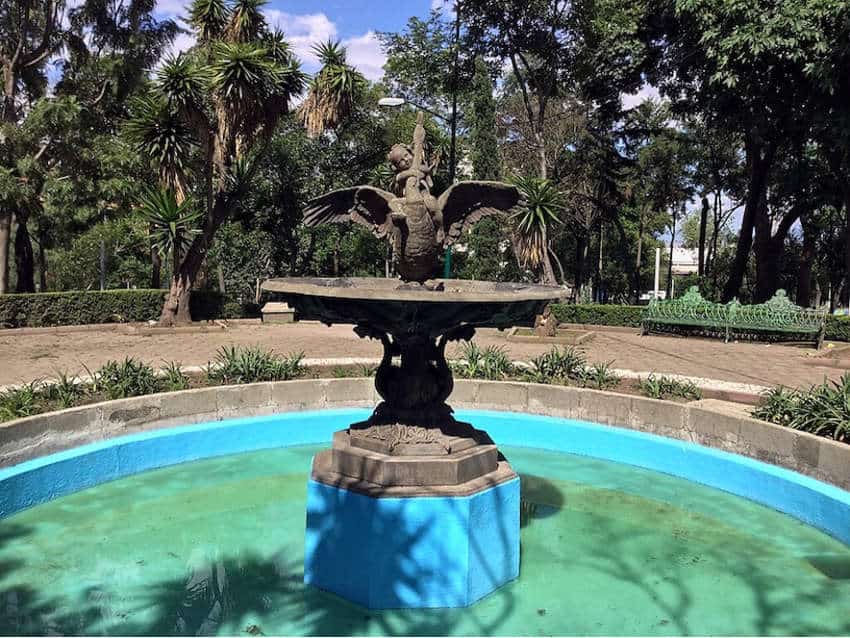 Una fuente adornada en Parque Lira en la Ciudad de México. Una escultura de piedra de un niño que monta un cisne se sienta en una base azul brillante dentro de una piscina pintada por turquesa, rodeada de bancos y árboles del parque.