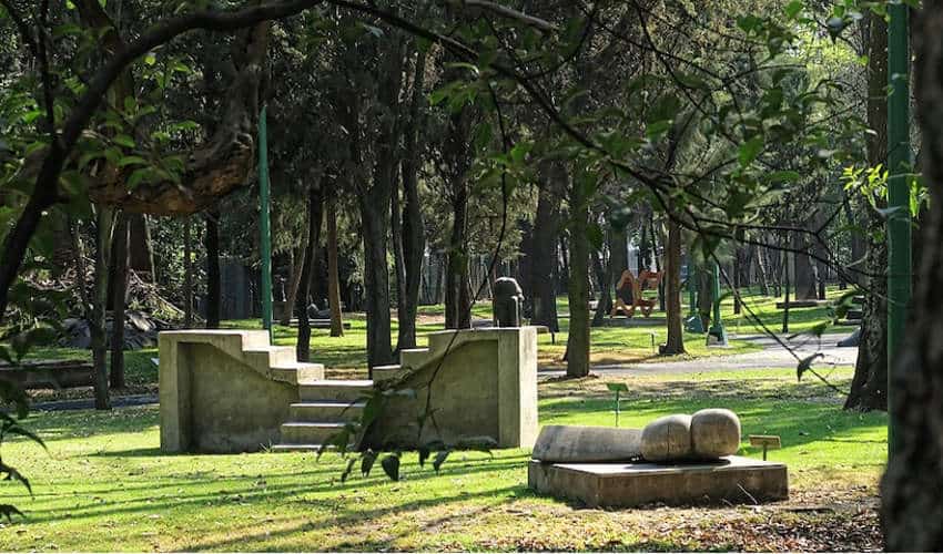 Una tranquila escena del parque con varias esculturas de arte moderno esparcidos por un césped verde, rodeada de árboles altos y luz solar moteada.