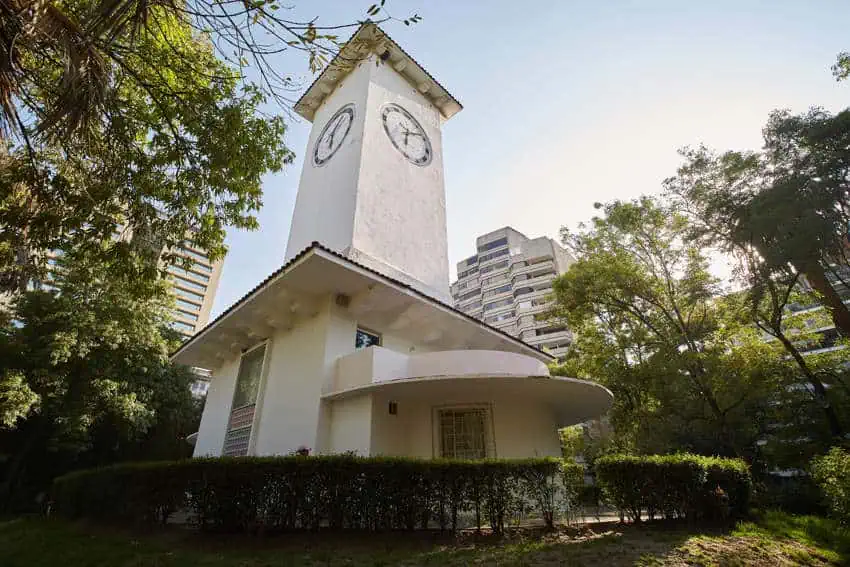 Una torre blanca de relojes art deco se ve desde un ángulo bajo en un día soleado en Parque Lincoln. En el fondo hay árboles circundantes y edificios modernos de la Ciudad de México.