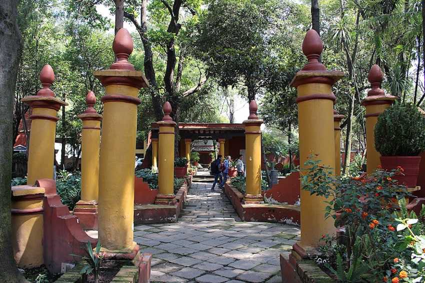 Un camino de piedra de piedra en los Jardines Nacionales de Fonoteca en la Ciudad de México. El camino está flanqueado por hileras de gruesos pilares de color amarillo mostaza con blusas decorativas de color marrón rojizo, todos ubicados dentro de una exuberante área boscosa verde.