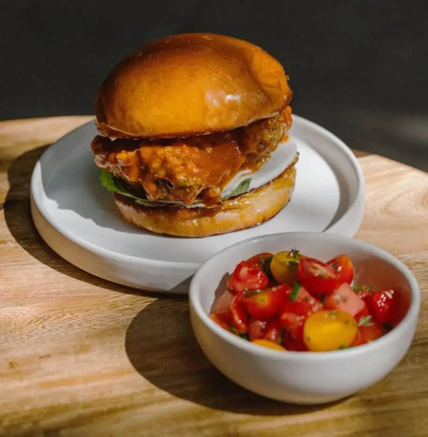 Un sándwich de pollo de aspecto jugoso con un moño dorado se encuentra en un plato blanco. Junto a él, un pequeño tazón blanco contiene una ensalada vibrante de tomates cherry rojos y amarillos. Ambos platos se colocan en una tabla de corte de madera clara.