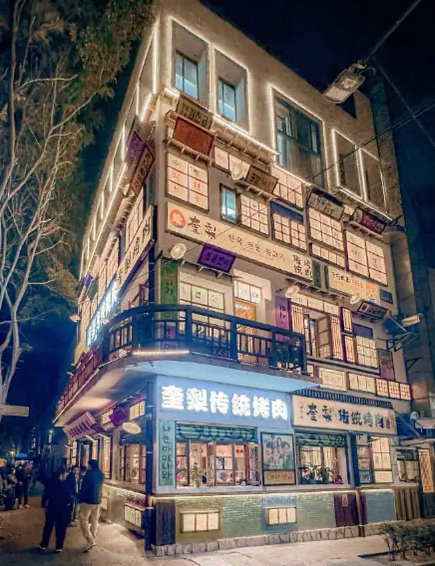 Un edificio de varios pisos se muestra por la noche. Está brillantemente iluminado con numerosas ventanas y letreros en su exterior. La gente camina por la acera frente al edificio. Los árboles en el lado izquierdo están desnudos, lo que sugiere que es un invierno o una temporada más fría.