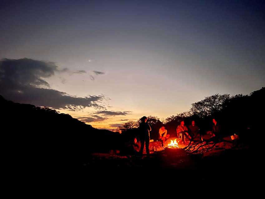 Un grupo de amigos se sienta alrededor de una fogata al anochecer en un campo en el estado de Aguascalientes, México. El sol ajustado ilumina el horizonte con un cálido brillo de naranja contra un cielo oscuro.