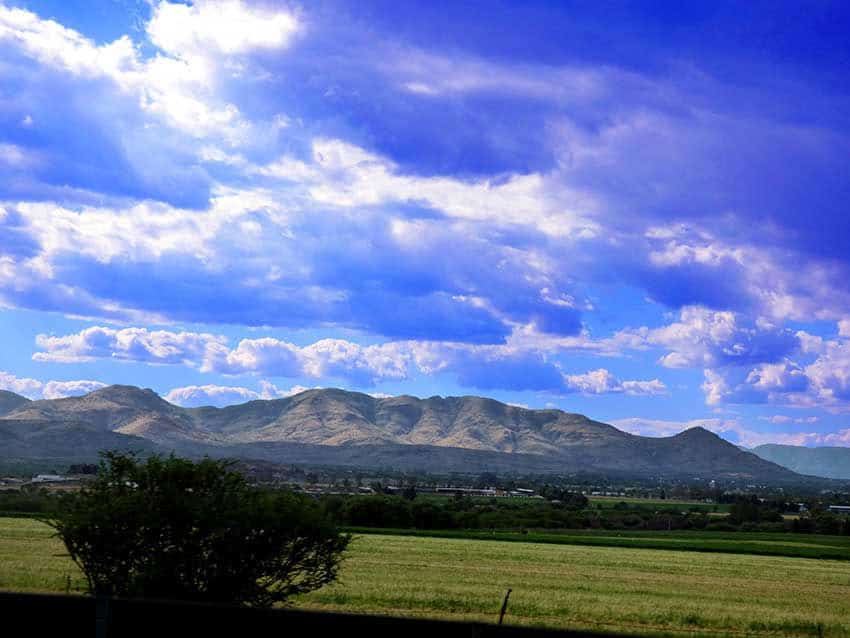 Una vista panorámica de la cordillera Cerro del Muerto en Aguascalientes, México, con un campo verde en primer plano bajo un vibrante cielo azul con nubes dispersas.