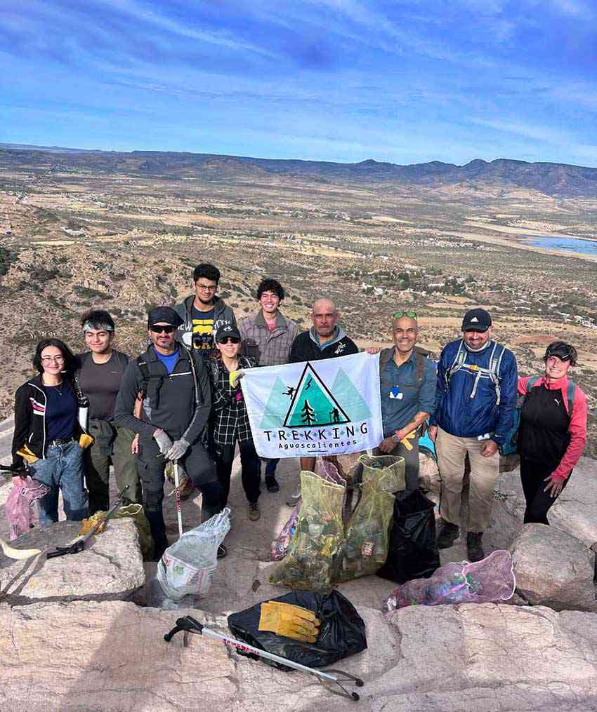 Un grupo de personas se encuentra en una cumbre rocosa, sosteniendo una bandera que dice "trekking aguascalientes". Están rodeados de bolsas de basura que han recolectado, con un paisaje pintoresco y árido y montañas visibles detrás de ellas.