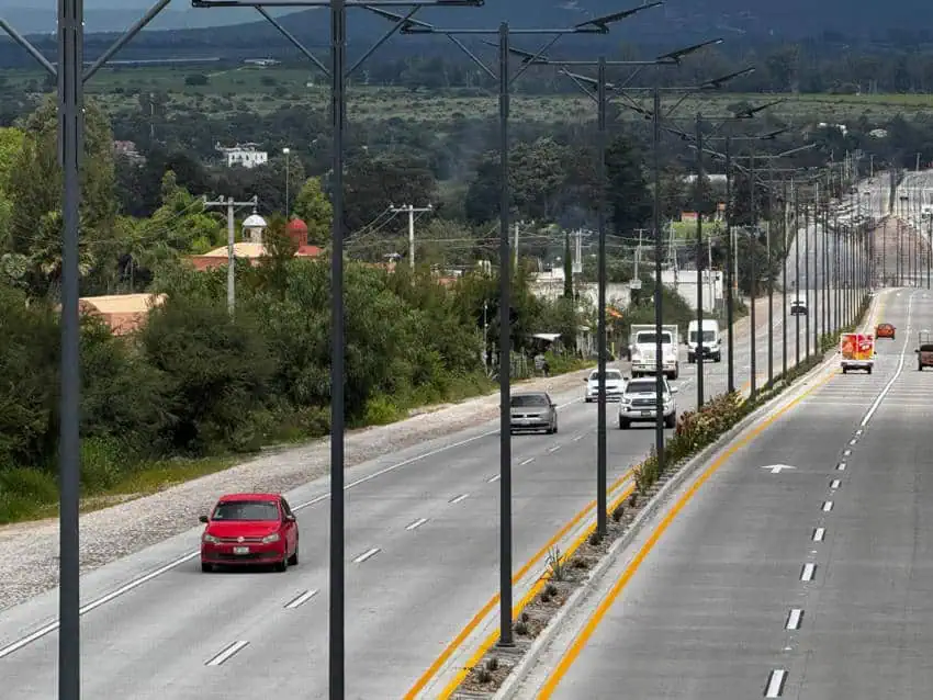 Una carretera de varios carriles que atraviesa un pequeño México. La carretera recientemente modernizada presenta pavimento fresco, una mediana ajardinada y una larga fila de nuevas farolas con energía solar. El camino conecta San Miguel de Allende y Dolores Hidalgo en México.