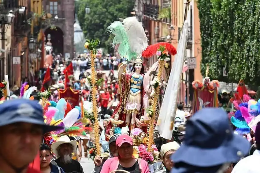 Una vista a nivel de la calle de una procesión religiosa abarrotada para el día de la fiesta de San Miguel Arcángel en San Miguel de Allende, México. Una estatua del santo patrón se lleva a una plataforma, rodeada de participantes en el desfile y las tradicionales y coloridas decoraciones de plumas.