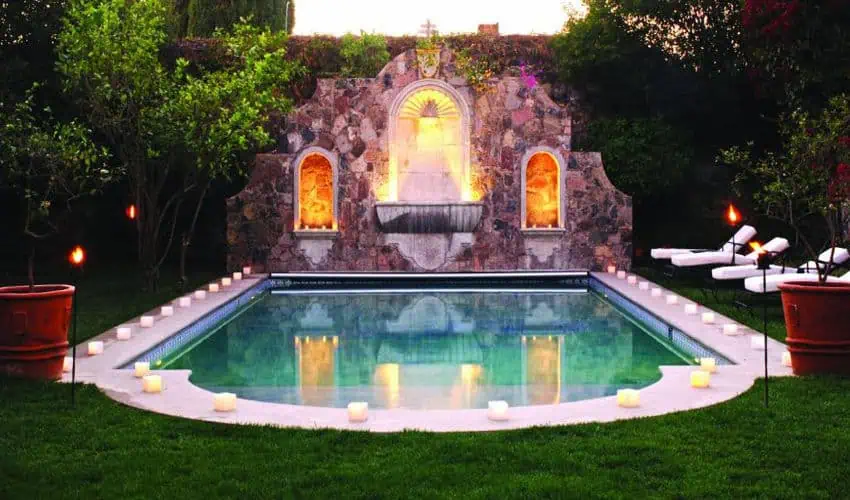 Una piscina a la luz de las velas en un hotel boutique de lujo en San Miguel de Allende, iluminado al anochecer con antorchas alrededor de la piscina. Un muro de piedra de estilo colonial con una fuente suavemente iluminada está en el fondo.