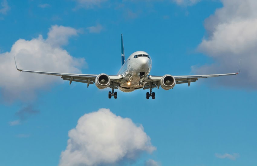 Un avión de pasajeros sin marcar de color plateado en un cielo azul con nubes hinchadas dispersas. El tren de aterrizaje del avión está abajo.