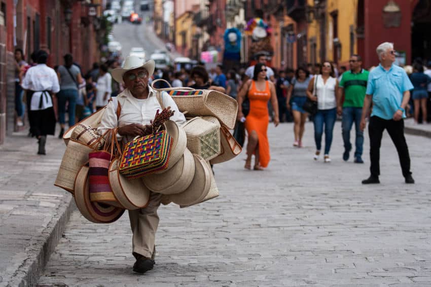 Un vendedor ambulante artesanal, con un sombrero tradicional de vaquero de paja, camina por una bulliciosa calle adoquinada en San Miguel de Allende. Lleva una carga de cestas de palma tejidas hechas a mano, sombreros y bolsas coloridas para la venta sobre sus hombros.