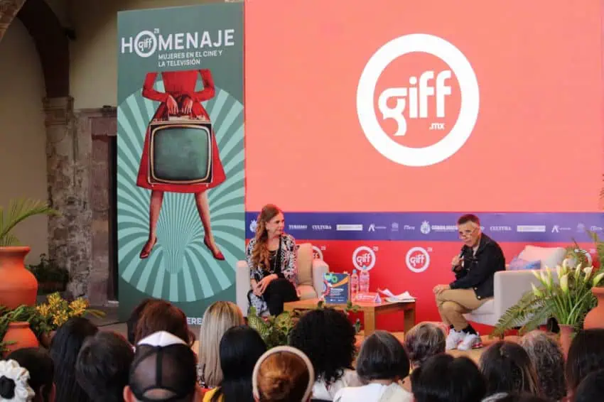 Una discusión en el panel en el escenario tiene lugar ante una audiencia en el Festival Internacional de Cine de Guanajuato (GIFF) en San Miguel de Allende, México. En el fondo, una pantalla grande muestra el logotipo del festival y una pancarta para su tributo especial, "HOMENAJE: Mujeres en el Cine y la televisión"