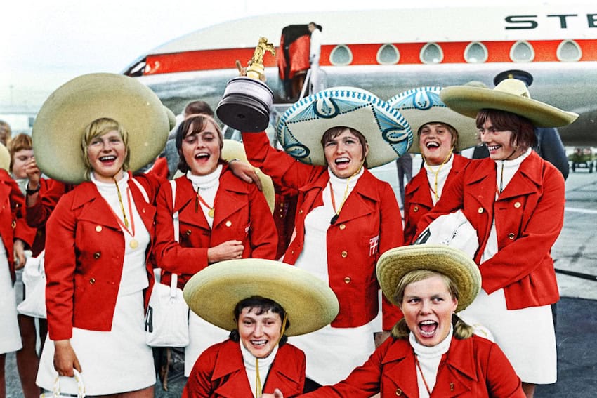 Un equipo de mujeres en chaquetas y sombreros rojos eleva el trofeo de la Copa Mundial en los escalones en un avión.
