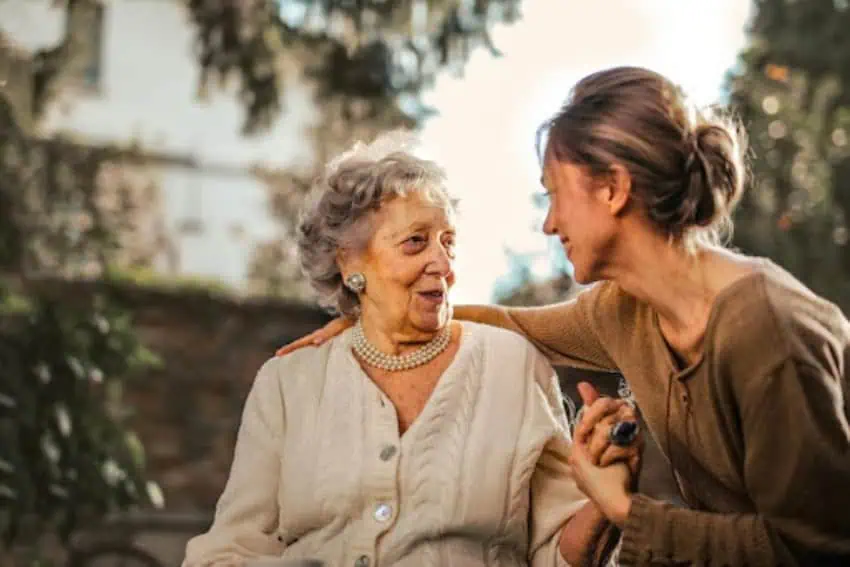 A caring younger woman provides support and companionship to an elegant elderly woman in a garden setting.