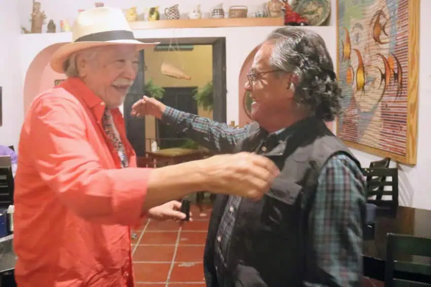 Dos mayores mexicanos se meten calurosamente con sonrisas y brazos abiertos dentro de una casa o estudio llenos de arte y cerámica.
