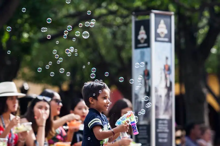 Un niño sonríe mientras juega con una pistola de burbujas en una plaza pública soleada en San Miguel de Allende. Está rodeado de adultos sentados en bancos de parques en el fondo.