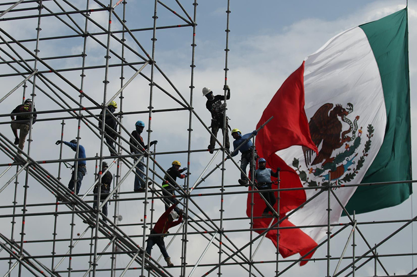 Workers install scaffolding in front of a giant Mexican flag