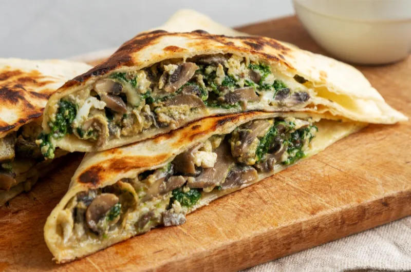 Un primer plano de una quesadilla de mitad de mitad, plegada y frita que descansa sobre una tabla de corte de madera. El relleno visible dentro de la tortilla de harina es una mezcla sabrosa de hongos marrones en rodajas (Champiñones), Epazote de hoja verde y queso blanco derretido.