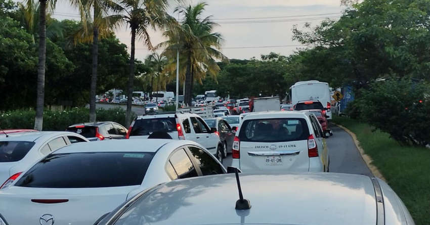 Una foto tomada desde el interior de un automóvil en tráfico pesado y lento en una carretera en Puerto Vallarta, Jalisco, México, que muestra una larga línea de autos, que incluyen una ambulancia y un autobús, parachoques a parachoques. El camino está lleno de exuberantes follaje tropical verde y palmeras debajo de un cielo nublado.