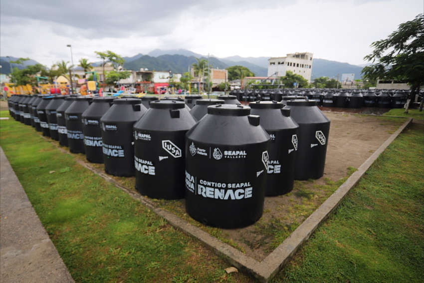 Una foto de ángulo bajo muestra docenas de nuevos tanques de almacenamiento de agua negra (probables rototas o cisternas similares) dispuestas en filas en un campo cubierto de hierba en Puerto Vallarta, Jalisco, México. Los tanques están marcados con logotipos para SeaPal Vallarta y el texto "Contigo Seapal Renace" (con usted, SeaPal renace). En el fondo hay edificios y montañas verdes bajo un cielo nublado.