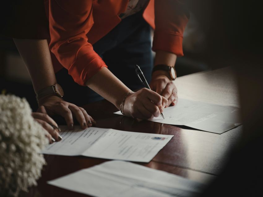 Una vista de las manos de una mujer firmando documentos colocados sobre una mesa de conferencias de madera. Un documento tiene lo que parece un escudo gubernamental de un país indeterminado.
