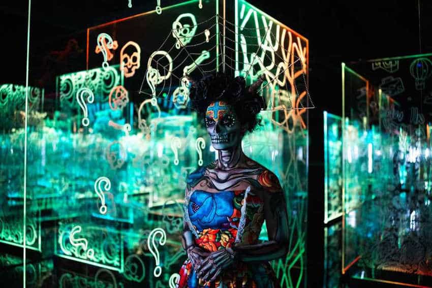 Una persona con un elaborado maquillaje de calavera y pintura corporal colorida se encuentra dentro de una instalación de arte moderno e inmersivo de paneles acrílicos de color verde brillante.