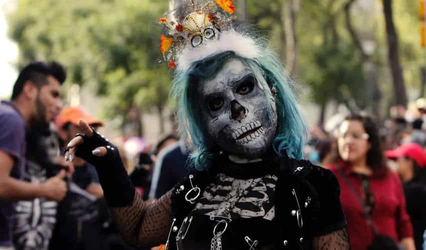 Persona con cabello verde azulado y una elaborada pintura facial de calavera posa para la cámara durante una celebración del Día de los Muertos, vistiendo un traje de estilo gótico y una pequeña corona con calaveras y caléndulas.