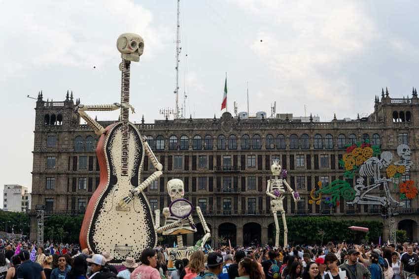 Figuras de esqueletos gigantes (calacas), incluida una con forma de guitarra, se elevan sobre una gran multitud en la plaza del Zócalo de la Ciudad de México, con el Palacio Nacional al fondo, durante la celebración del Día de Muertos.