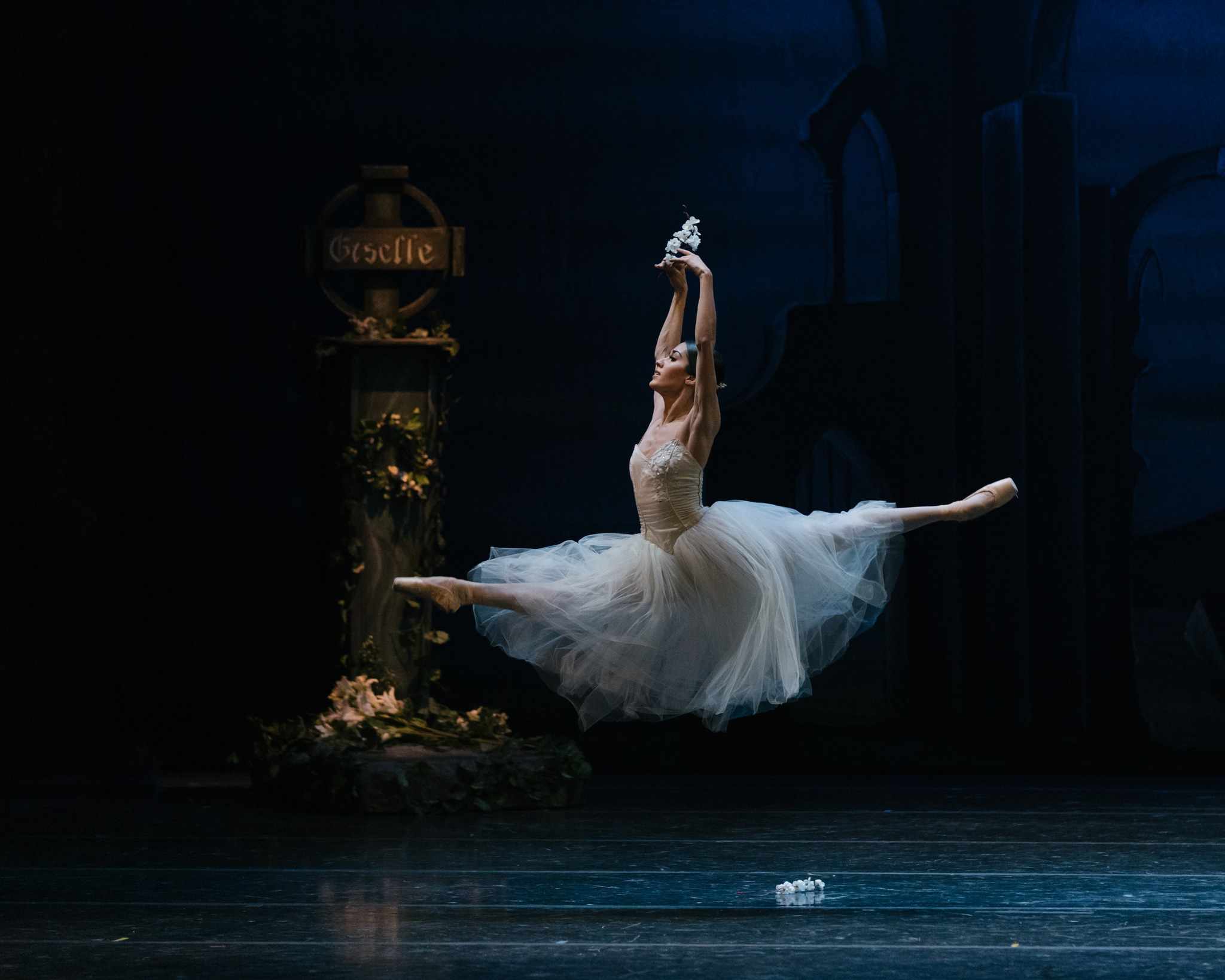 Una bailarina con un tutú blanco realiza un gran jeté en un escenario oscuro, con una lápida marcada "Giselle" visible al fondo.