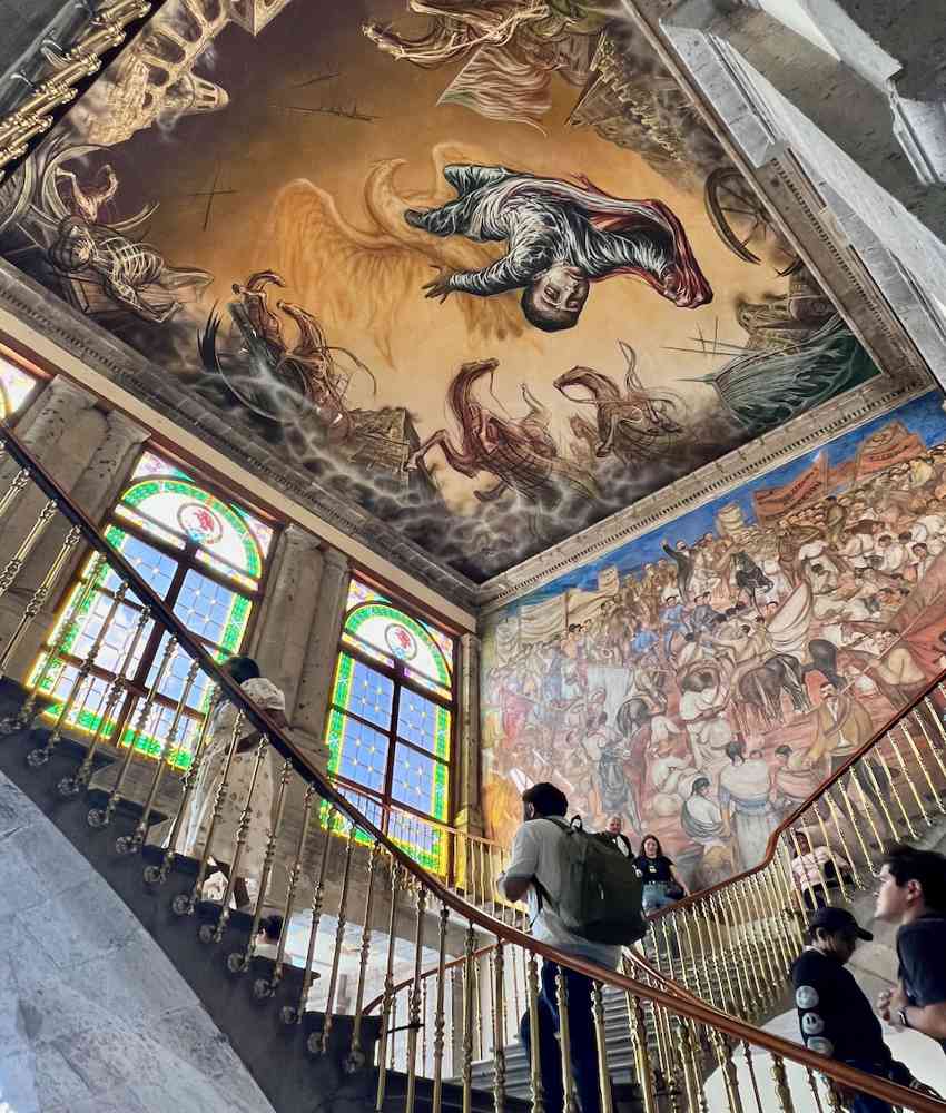 Una amplia y ornamentada escalera de piedra dentro del Castillo de Chapultepec muestra a los visitantes subiendo las escaleras, rodeados por un espectacular fresco en el techo que representa la conquista de México por los españoles, así como un mural en la pared al lado de las escaleras.