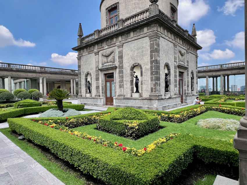Los jardines bellamente cuidados en el laberinto de la azotea del Castillo de Chapultepec, con setos simétricos de boj, parterres de flores y un pequeño edificio de ladrillos de cantera de estilo barroco en el centro de la foto. El edificio está decorado con estatuas.