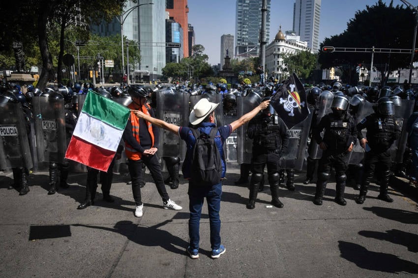 Un manifestante solitario con un sombrero de vaquero y un chaleco naranja se encuentra con los brazos extendidos, sosteniendo una bandera mexicana y otra bandera con una calavera y tibias cruzadas, frente a una fila de policías antidisturbios mexicanos con cascos y escudos en una calle soleada de la ciudad.