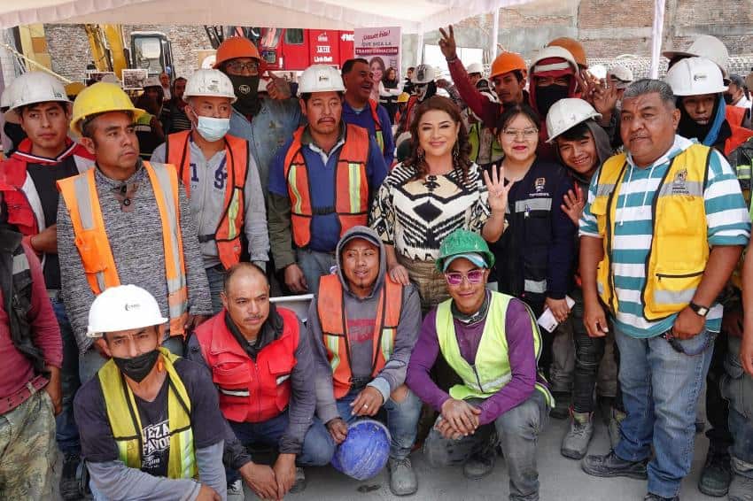 Un grupo de trabajadores de la construcción con chalecos amarillos y naranjas y cascos posando para una foto grupal con dos mujeres políticas.