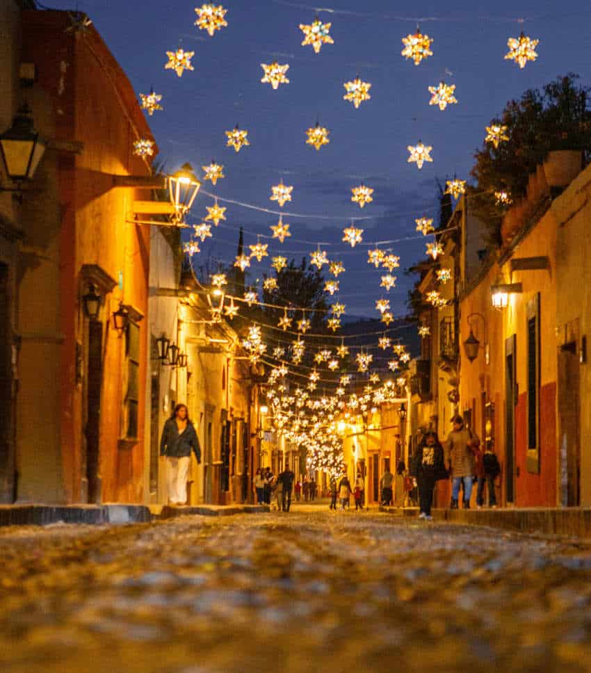 Una vibrante toma en ángulo bajo de una calle estrecha de adoquines en un pueblo mexicano al anochecer, iluminada por numerosas luces doradas en forma de estrellas colgadas entre edificios coloridos, con gente paseando por la calle.