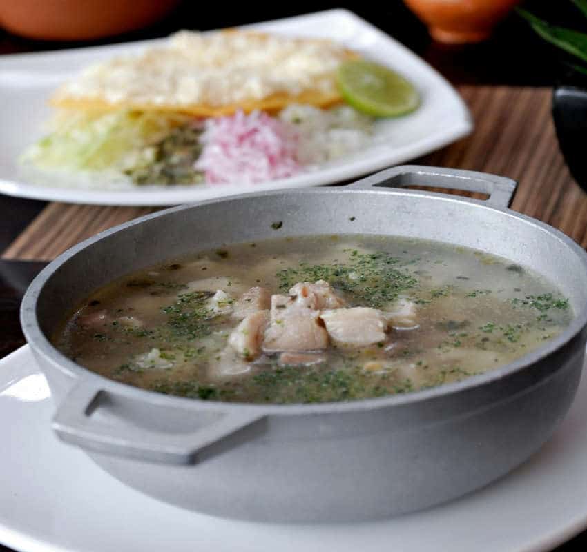 Un tazón de metal lleno de pozole blanco tradicional (caldo claro, trozos de pollo o cerdo y hierbas) servido junto con una guarnición, perfecto para una celebración del Día de Muertos.