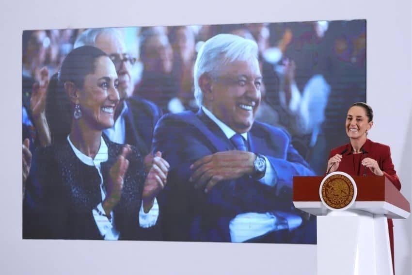 Claudia Sheinbaum en el podio presidencial frente a una gran proyección de una fotografía de ella y Andrés Manuel López Obrador sonriendo entre el público mientras observan un evento.