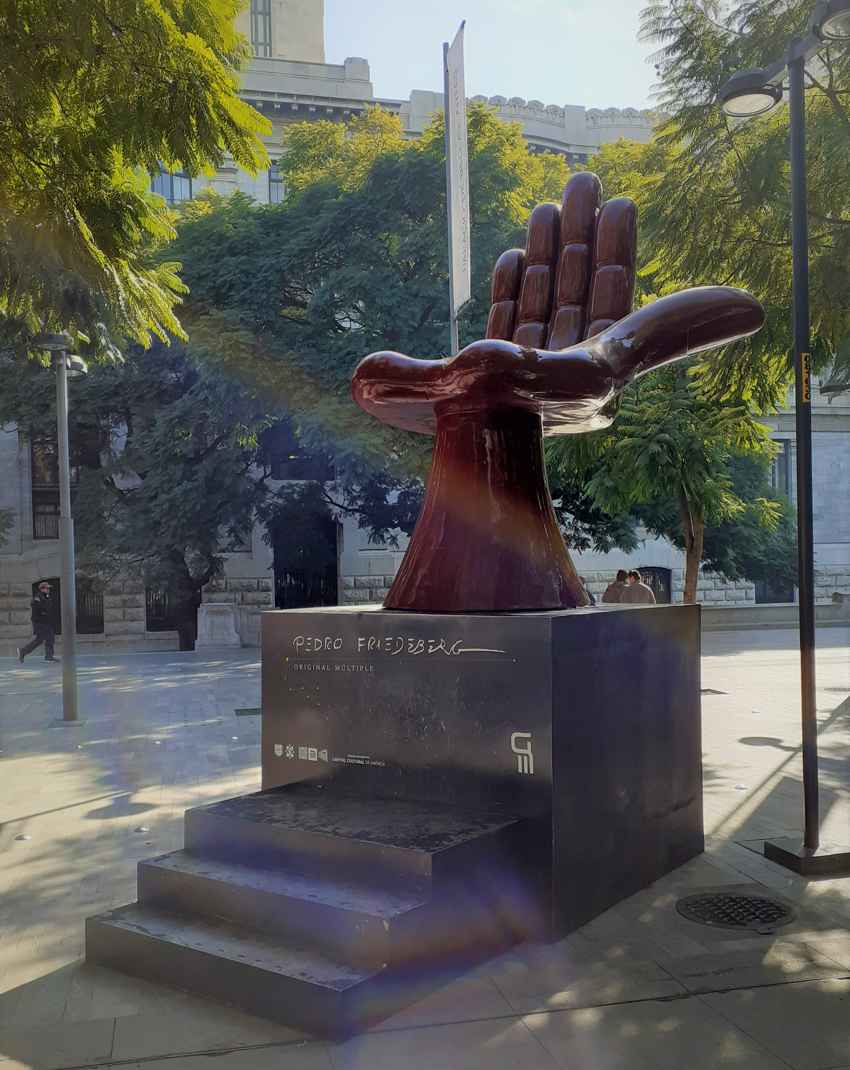 Una monumental escultura de Silla de Mano de color marrón rojizo del artista mexicano Pedro Friedeberg se asienta sobre un pedestal escalonado negro en el parque Alameda Central de la Ciudad de México, enmarcada por árboles verdes y arquitectura histórica.