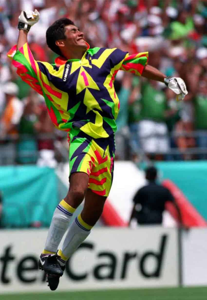 El futbolista mexicano Jorge Campos en un partido del Mundial de 1994. Está atrapado en medio de un alegre salto con un puño en el aire, vistiendo un uniforme de color verde neón, rosa neón, amarillo y azul. y calcetines blancos, azules y amarillos.