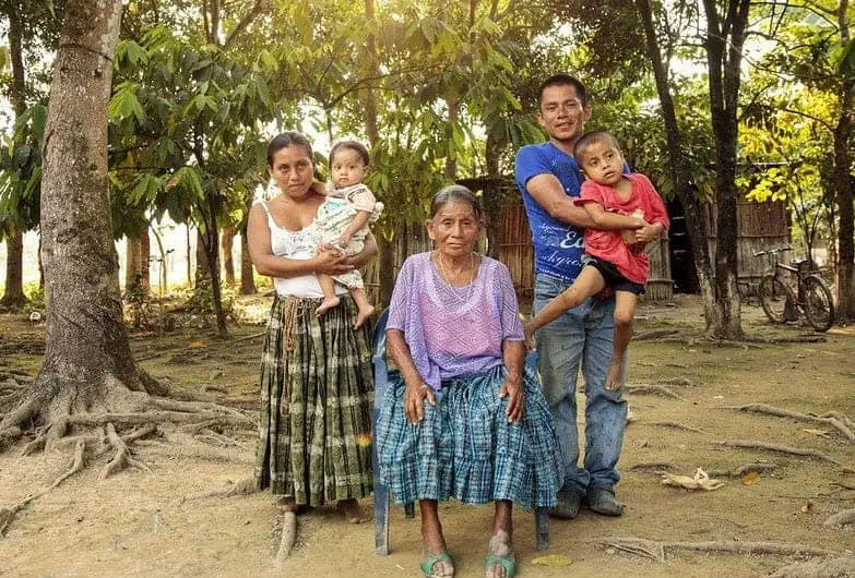 Una familia indígena maya posa al aire libre. La madre y el padre están de pie, cada uno con un niño en brazos, mientras la abuela permanece sentada.