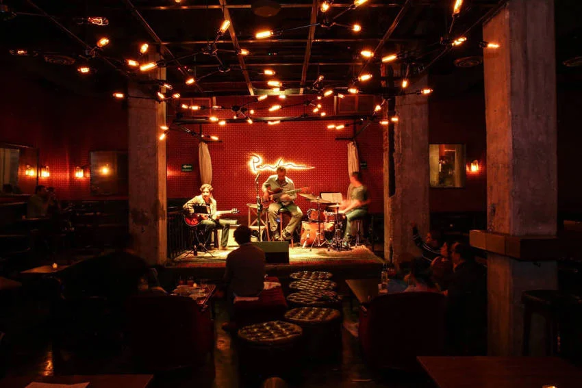 Un trío de jazz se presenta en el escenario bajo luces cálidas y brillantes para una audiencia en Parker & Lenox, un club de jazz íntimo de estilo clandestino en la Ciudad de México.