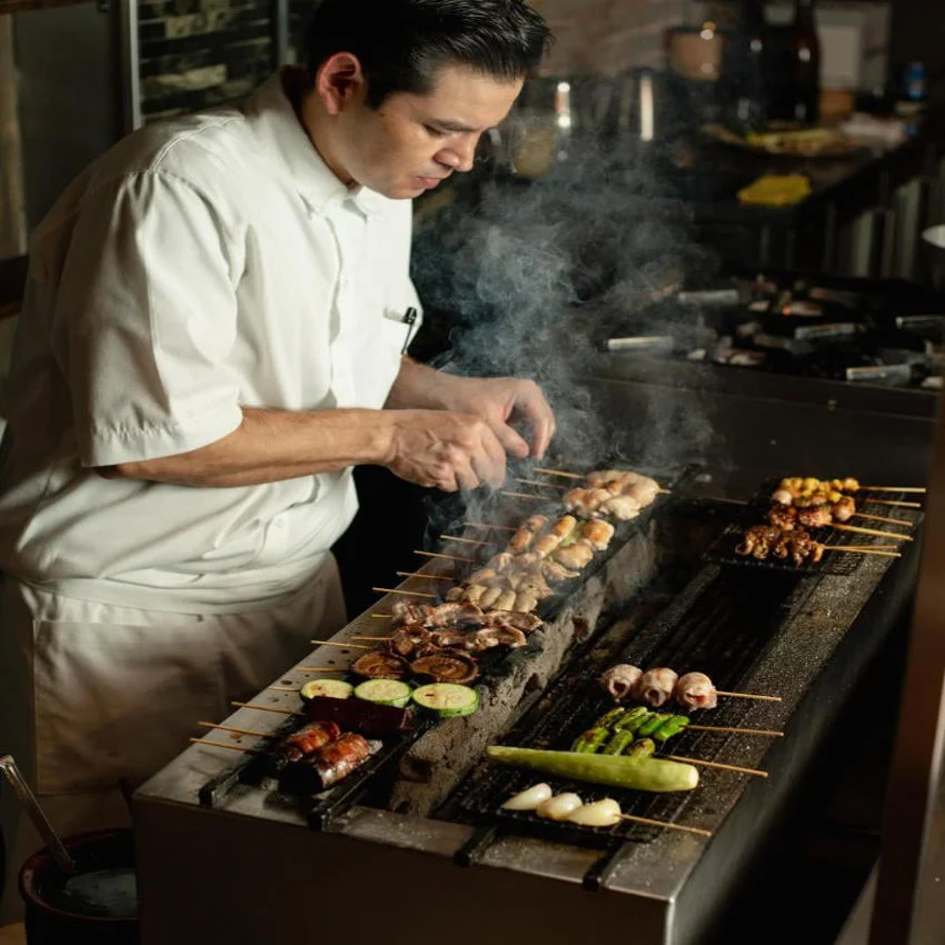 Un chef prepara yakitori japonés, asando brochetas de mariscos, carne y verduras en una parrilla robata en un elegante restaurante en México.