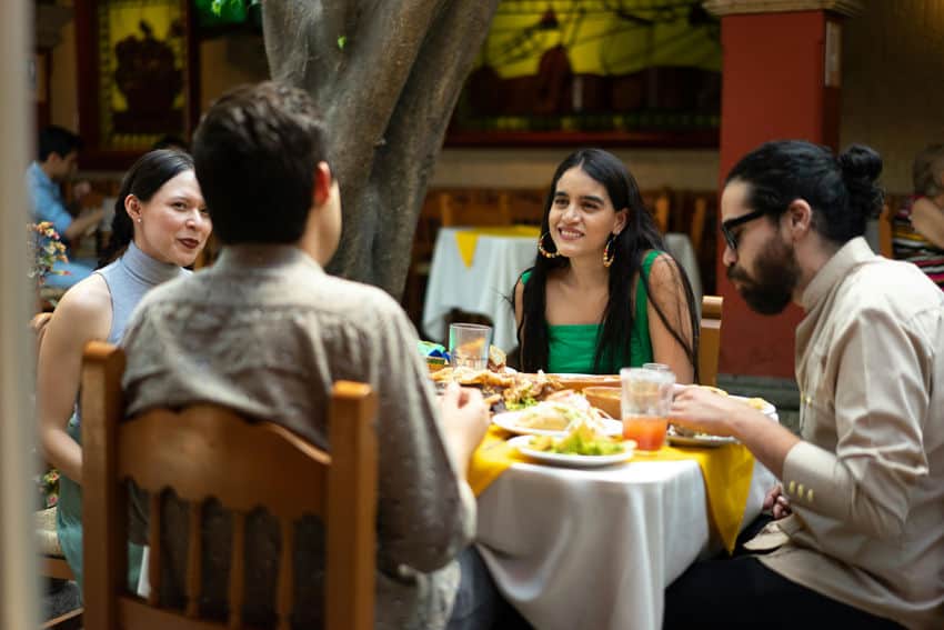 Dos hombres jóvenes y dos mujeres jóvenes cenan y conversan alrededor de una mesa de restaurante con comida mexicana en un luminoso restaurante con patio a la sombra de los árboles.