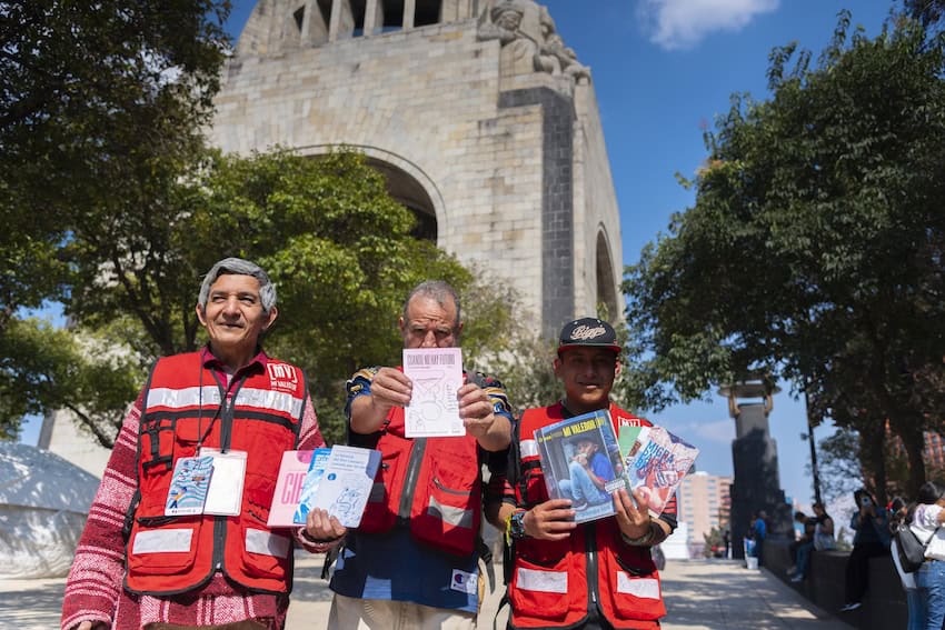 Un grupo de 3 hombres con chalecos rojos vendiendo un periódico callejero en la Ciudad de México.